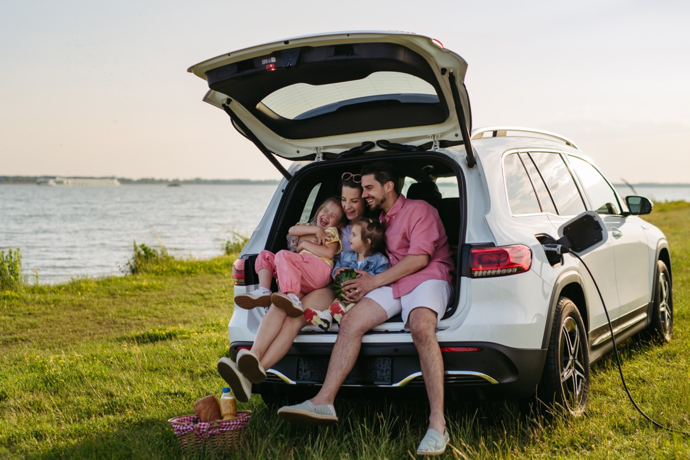 Familie på fire koser seg i bagasjerommet på bilen, parkert ved ein innsjø med ein piknikkurv ved sida av.
