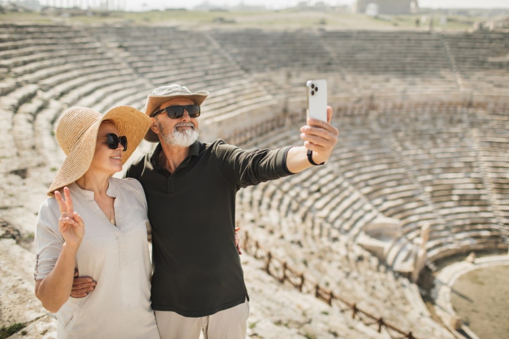 Eldre par tek ein selfie framfor eit gamalt amfiteater, begge smilande med solhattar og solbriller.