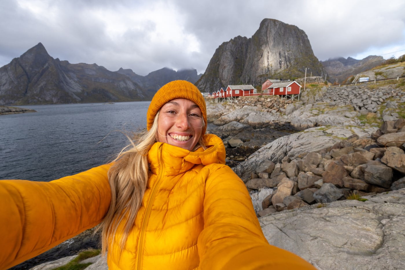 Strålande kvinne i gul jakke tek ein selfie med den majestetiske norske kysten og raude hytter i bakgrunnen, illustrerer varme og glede i enkelheita ved livet.