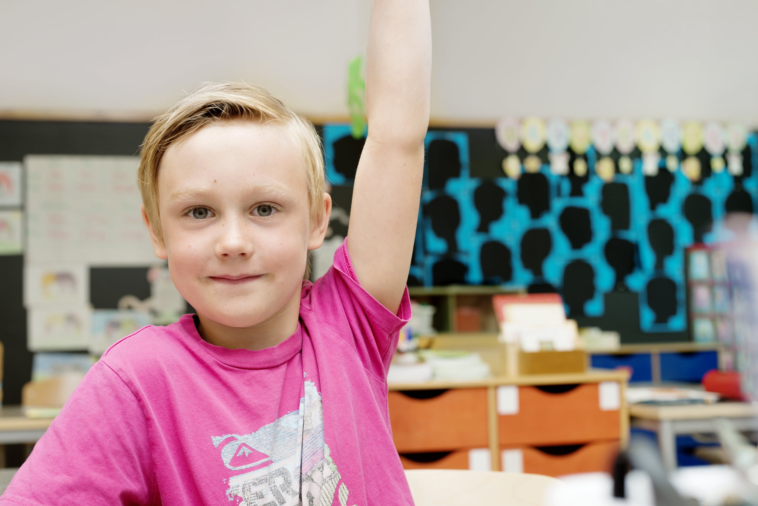 Skulegut i rosa T-skjorte som rekk opp handa i eit klasserom.