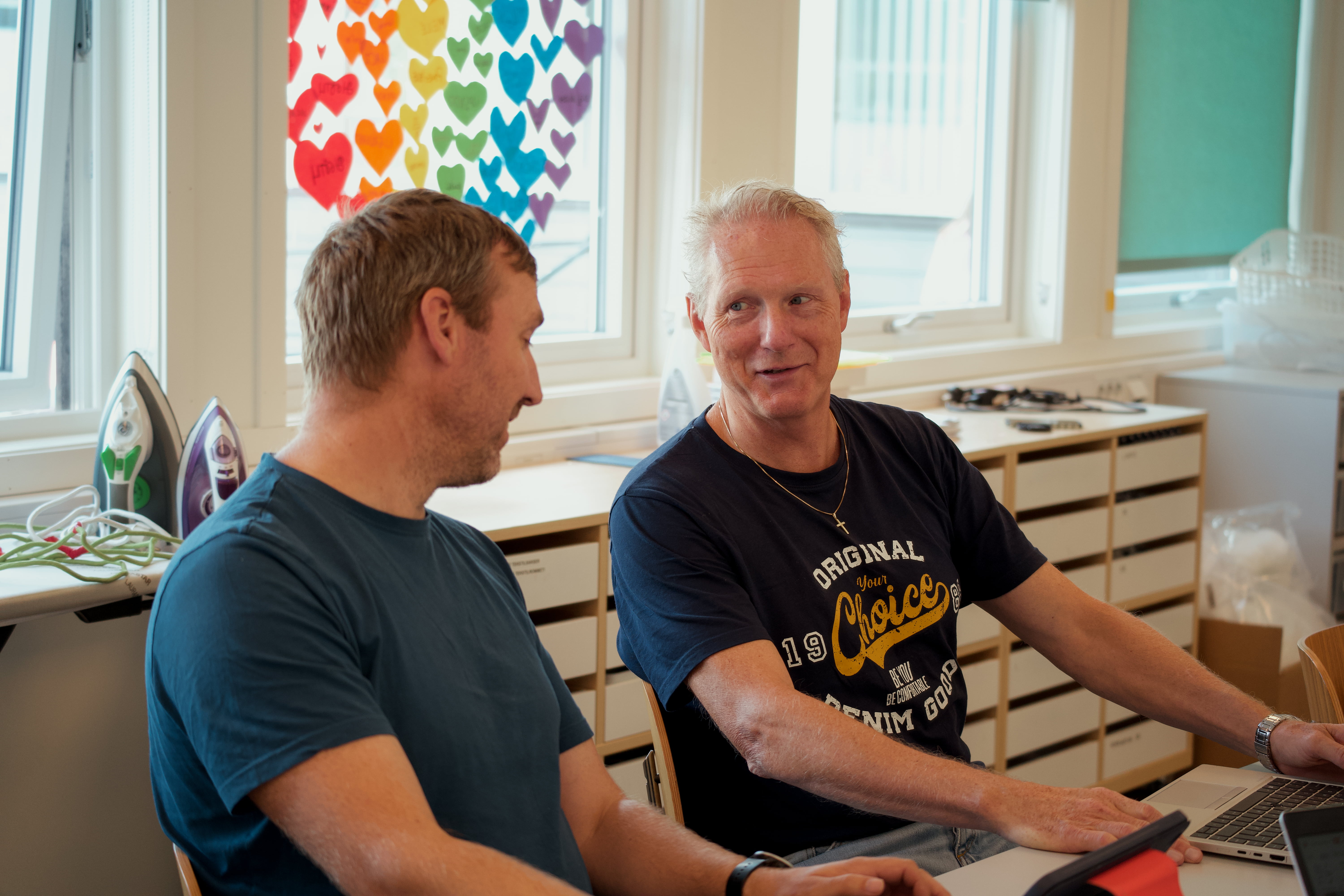 To personar sit ved eit bord og jobbar på ein berbar PC i eit rom med store vindauge. På veggen heng ein plakat med fargerike hjarter, og det står strykejarn på ei hylle bak dei.