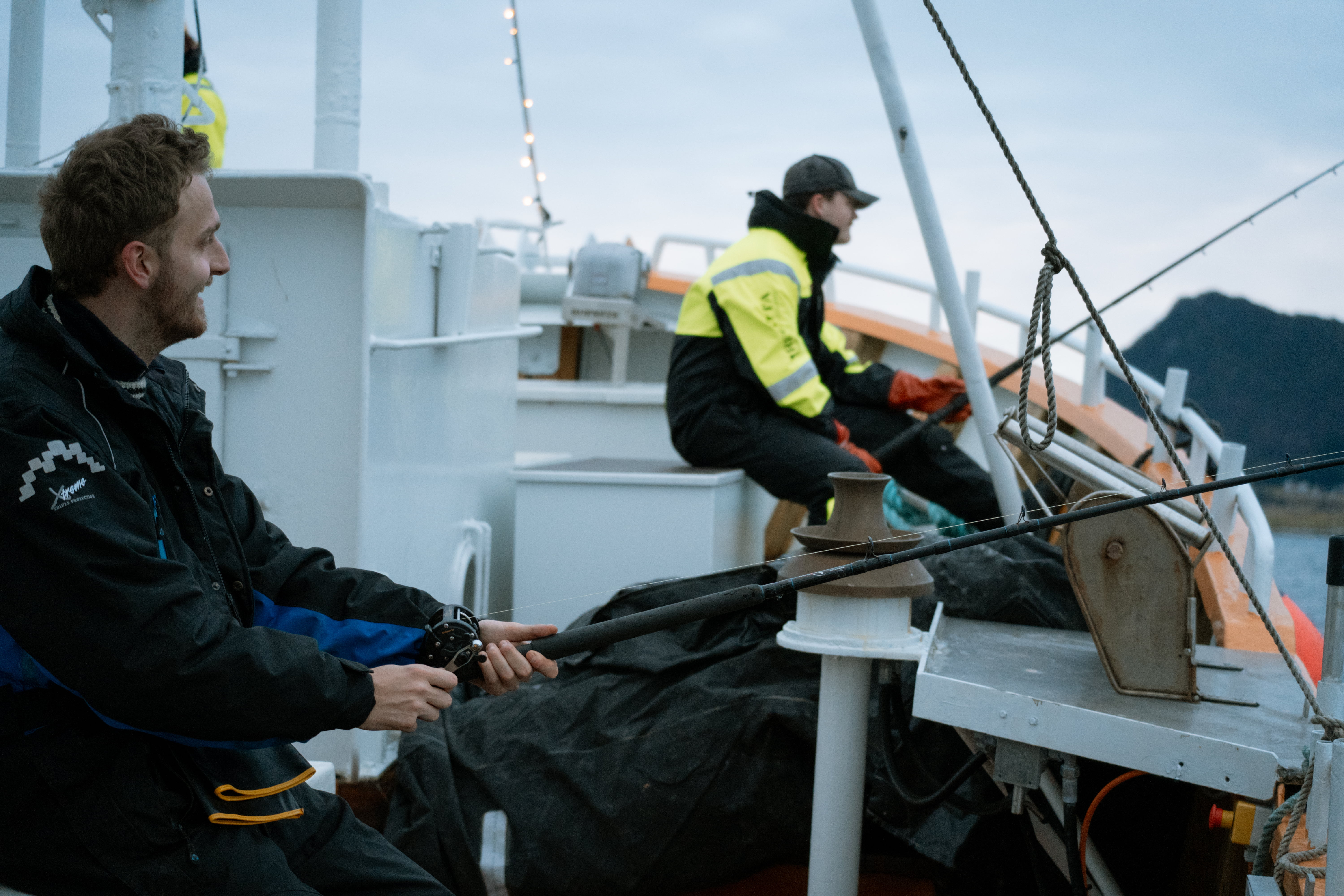 To personar fiskar frå ein båt med fjell i bakgrunnen.