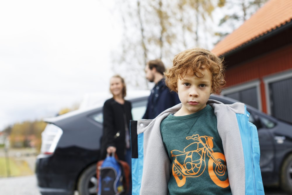 Liten gut med ryggsekk ser tankefullt ut medan han står ved sidan av ein svart bil, med familien i bakgrunnen.