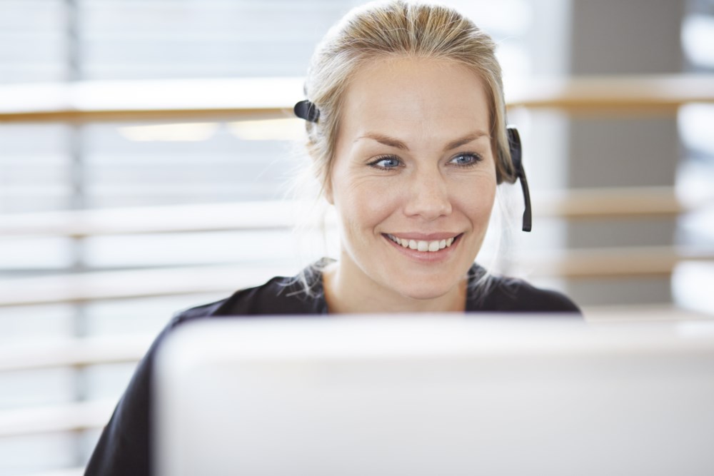 Ei blid kvinne med headset jobbar på datamaskin, ser engasjert og lyttande ut i ein lys og moderne kontorsetting.