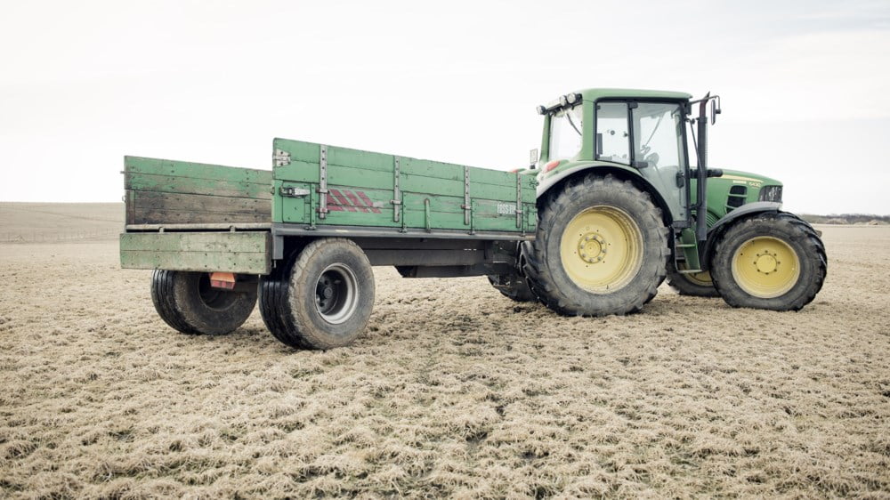 Grøn traktor med tilhengar står på eit ope jorde under ein lys himmel.